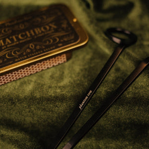 Detail shot of vintage-style gold tin matchbox and matte black wick trimmer engraved with Phantom Row.
