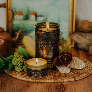 Lifestyle top shot of lit Cottage by the Sea 15 oz soy candle with a vintage green glass jar, cork top, and 3.3 oz gold tin candle arranged with shells, florals, and painting of the seaside.