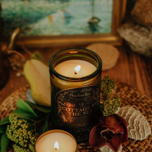 Lifestyle top shot of lit Cottage by the Sea 15 oz soy candle with a vintage green glass jar, cork top, and 3.3 oz gold tin candle arranged with shells, florals, and painting of the seaside.