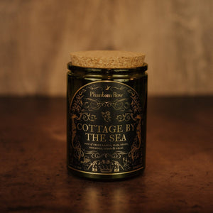 Front shot of Cottage by the Sea 11 oz soy candle with a vintage green glass jar, cork top, and black and gold foil label with vintage illustrations.