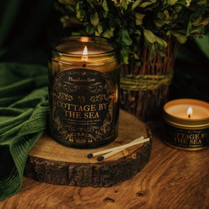 Lifestyle shot of lit Cottage by the Sea 11 oz soy candle with a vintage green glass jar, cork top, and 3.3 oz gold tin candle arranged on a wood stand with greenery and velvet.