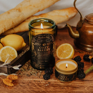Lifestyle shot of lit Cozy Bakery 15 oz soy candle with a vintage green glass jar, cork top, and 3.3 oz gold tin candle arranged with baguettes, lemons, blackberries, a copper tea kettle, and dried florals.