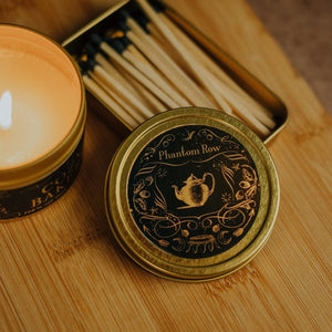 Detail shot of the Cozy Bakery 3.3 oz gold tin candle lid with black and gold foil detail propped on top of a gold mathbox tin with black-tipped matches and a lit tin candle nearby.
