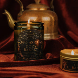 Lifestyle shot of lit Cozy Bakery 11 oz soy candle with a vintage green glass jar, cork top, and 3.3 oz gold tin candle arranged on red velvet with a copper kettle in the background.