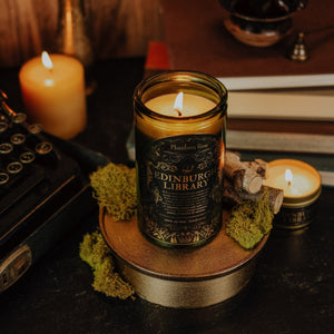 Lifestyle shot of lit Edinburgh Library 15 oz soy candle with a vintage green glass jar, cork top, and 3.3 oz gold tin candle arranged with a stack of books, vintage typewriter, and moss.