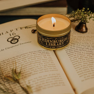Lit Edinburgh Library 3.3 oz gold tin soy candle sitting on top of open book arranged with a bronze candle snuffer and dried florals.