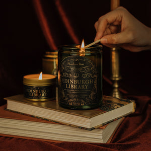 Lifestyle shot of lit Edinburgh Library 11 oz soy candle with a vintage green glass jar, cork top, and 3.3 oz gold tin candle arranged on a stack of book with a brass candlestick holder in the background.