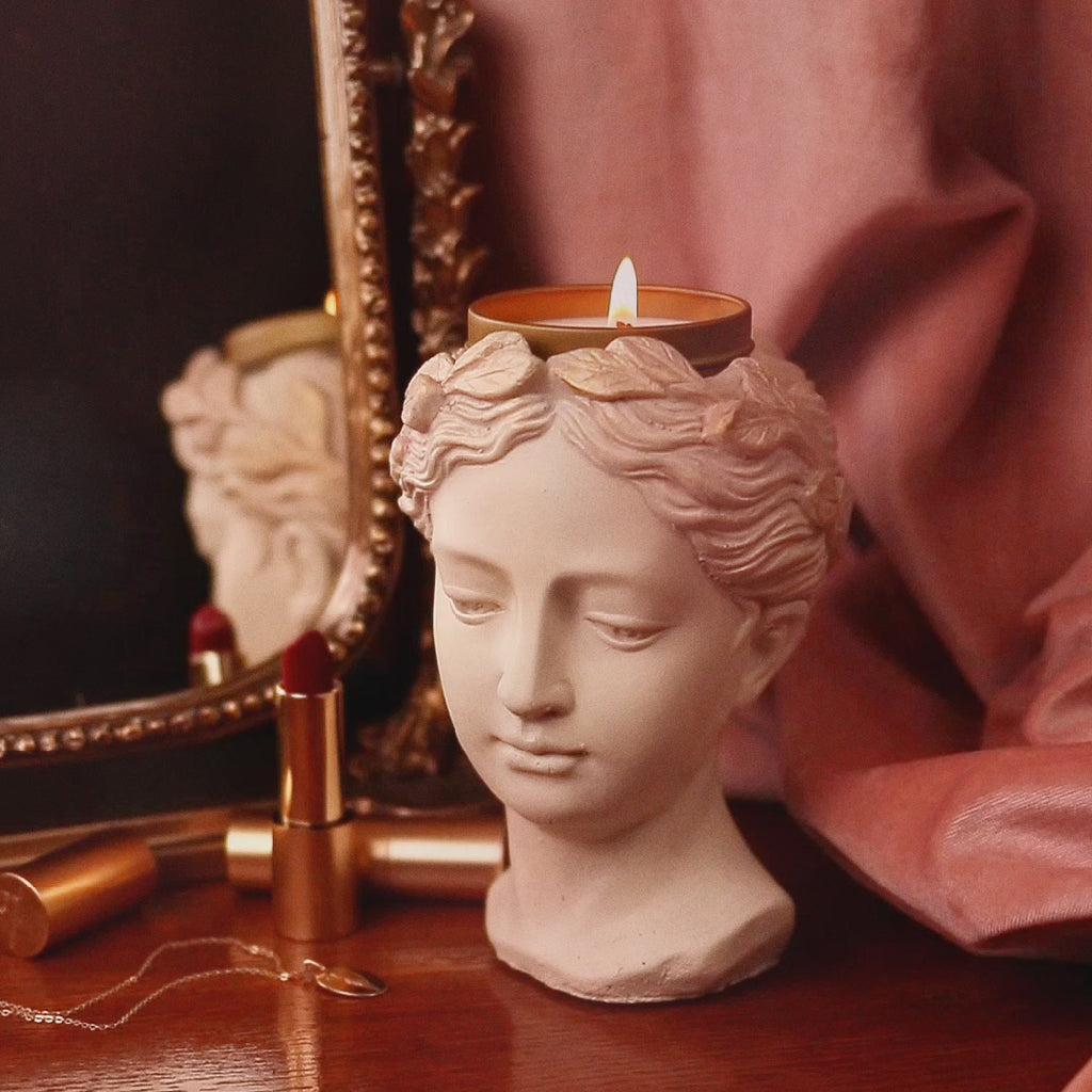 Gif showing handmade concrete greek goddess bust with gold candle tin in her crown sitting on vanity next to a mirror, lipstick, and jewelry,, and then a hand replaces candle with various crystals.