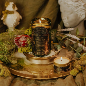 Lifestyle shot of lit Fairy Springs 15 oz soy candle with a vintage green glass jar, cork top, and 3.3 oz gold tin candle arranged on a reflective gold platter filled with clear water and bubbles, and surrounded by green dried florals and statues.