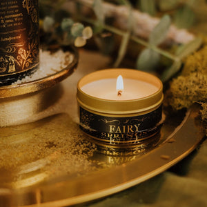 Lit Fairy Springs 3.3 oz gold tin soy candle sitting in a round gold tray filled with salt and water, reflecting the candle label in the water.