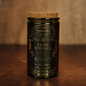 Front shot of Fairy Springs 15 oz soy candle with a vintage green glass jar, cork top, and black and gold foil label with vintage illustrations.