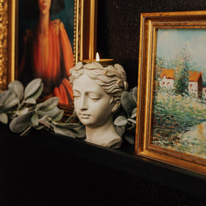 Handmade concrete goddess bust with lit gold tin soy candle in her crown sitting on a black picture ledge between two paintings with gold frames and a garland of greenery behind the statue.