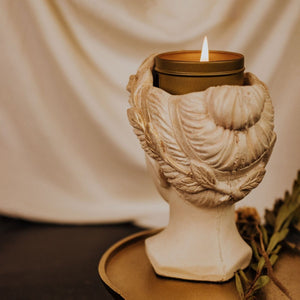 Back view of handmade concrete goddess bust statue, hand painted in ivory color with gold accents in her hair and leaf crown with lit gold tin candle sitting in her crown.