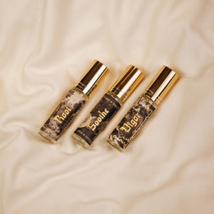 Hand holding a trio of Phantom Row mini-sized Ritual Mists in 0.3 oz glass atomizer vials with gold lids arranged on cream fabric. Root, Vigor, and Soothe labels show black and white vintage illustrations with the names in gold foil.