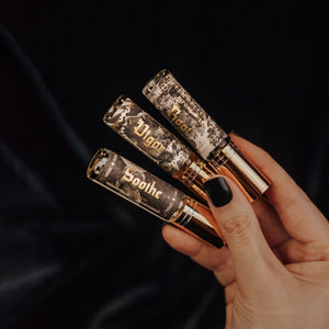 Hand holding a trio of Phantom Row mini-sized Ritual Mists in 0.3 oz glass atomizer vials with gold lids. Root, Vigor, and Soothe labels show black and white vintage illustrations with the names in gold foil.