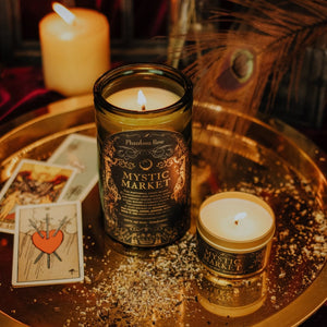 Lifestyle top shot of lit Mystic Market 15 oz soy candle with a vintage green glass jar, and 3.3 oz gold tin candle arranged on a reflective gold platter with tarot cards, spices, and an ostrich feather.