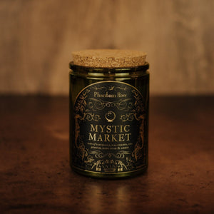 Front shot of Mystic Market 11 oz soy candle with a vintage green glass jar, cork top, and black and gold foil label with vintage illustrations