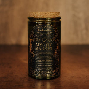 Front shot of Mystic Market 15 oz soy candle with a vintage green glass jar, cork top, and black and gold foil label with vintage illustrations