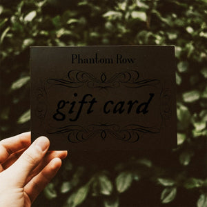 Hand holding up a black Phantom Row gift card with greenery blurred in the background.