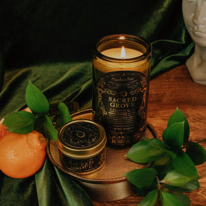 Lifestyle shot of lit Sacred Grove 15 oz soy candle with a vintage green glass jar, cork top, and 3.3 oz gold tin candle arranged on a gold stand with green leaves, an orange, concrete goddess bust, and green velvet.