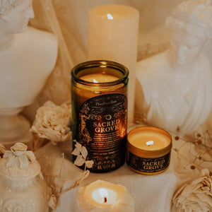 Overhead shot of lit Sacred Grove 15 oz soy candle with a vintage green glass jar, cork top, and 3.3 oz gold tin candle arranged on a ivory scene with tall ivory cylinder candle, ivory Apollo and Artemis bust statues, and cream-colored flowers.