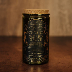 Front shot of Sacred Grove 15 oz soy candle with a vintage green glass jar, cork top, and black and gold foil label with vintage illustrations.
