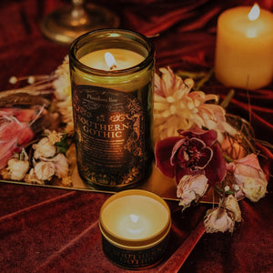 Lifestyle shot of lit Southern Gothic 15 oz soy candle with a vintage green glass jar, cork top, and 3.3 oz gold tin candle arranged with romantic flowers and sitting on dark red velvet.