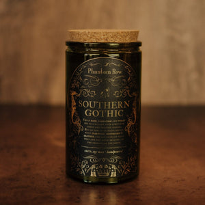 Front shot of Southern Gothic 15 oz soy candle with a vintage green glass jar, cork top, and black and gold foil label with vintage illustrations.