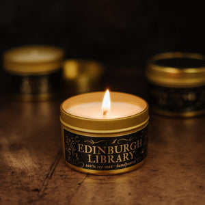 Close up of lit Edinburgh Library 3.3 oz gold soy candle tin with two blurred candle tins in the dark background.