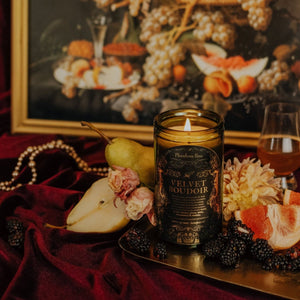 Lifestyle shot of lit Velvet Boudoir 15 oz soy candle with a vintage green glass jar,  arranged on a gold, rectangular platter with dried florals, pears, blackberries, a pearl necklace and a glass of amber liquid.
