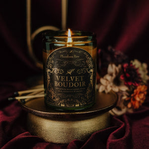 Lit Velvet Boudoir 11 oz soy candle sits on a gold stand with matches, florals, and a brass candlestick holder in the background with dark red velvet.