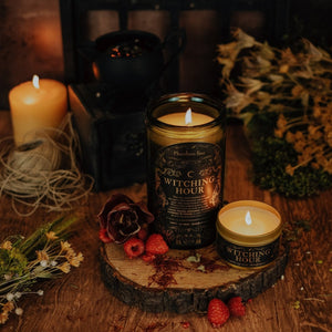Lifestyle shot of lit Witching Hour 15 oz soy candle with a vintage green glass jar,  and 3.3 oz golden candle tin arranged on a wood stump with raspberries, maroon flower, dried florals, and saffron spice, on a wood table.