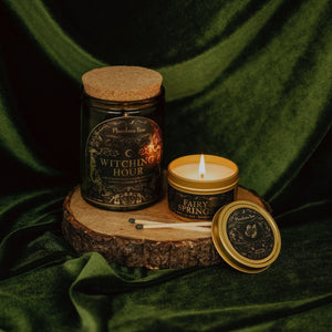 Lifestyle shot of lit Witching Hour 11 oz soy candle with a vintage green glass jar, cork top, and 3.3 oz gold tin candle arranged on a wood stand with a green velvet background.